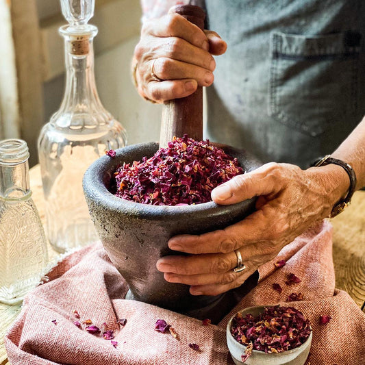 Dried red rose petals in a mortar while a woman uses a pestle to grind them finer.