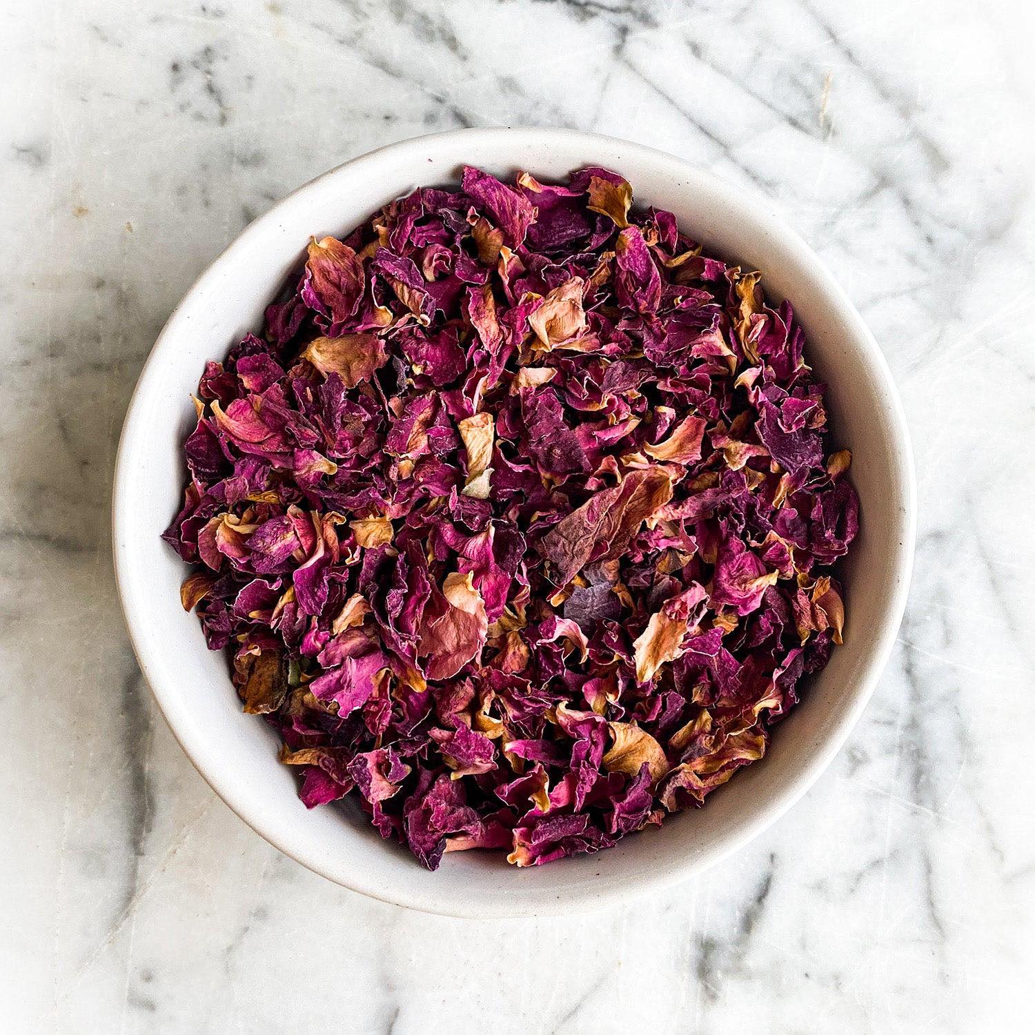 Dried red rose petals in a white bowl on a white marble table.