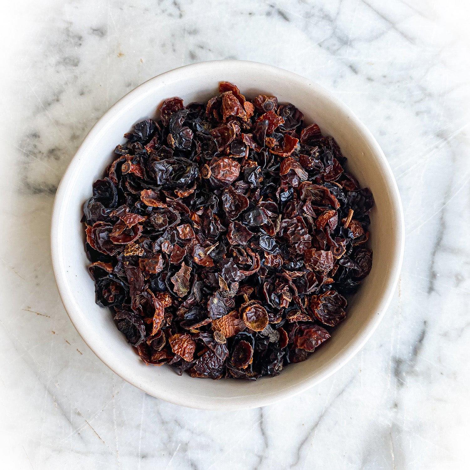 Dried rosehips in a white bowl on a white marble background.