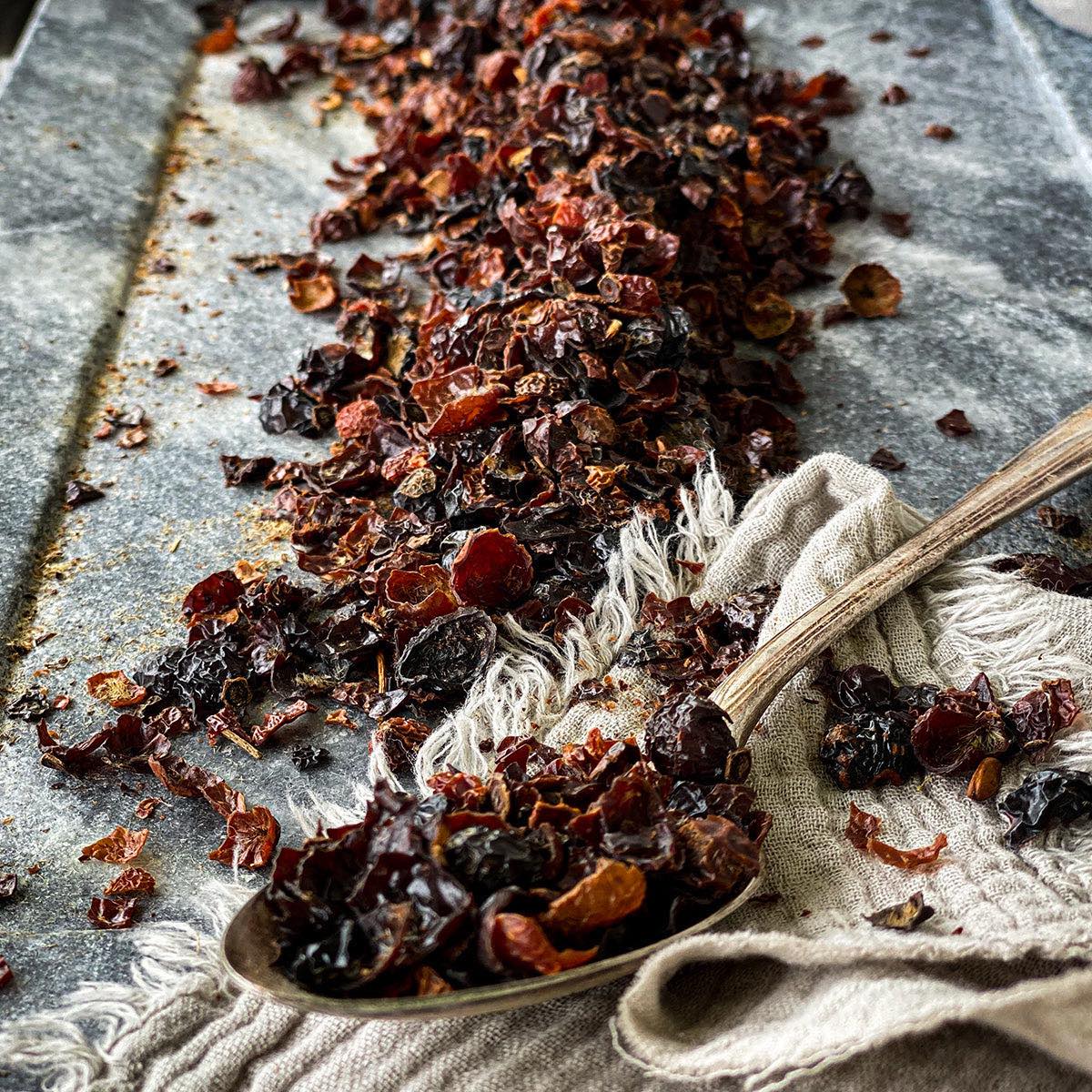 Dried rose hips on a metal background with a spoon filled with rosehips in the foreground.