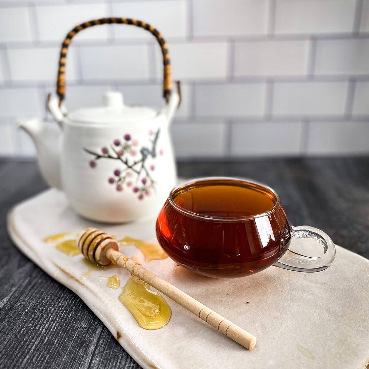 Dried rosehip tea in a clear mug with a honey wand and a white tea pot in the background.