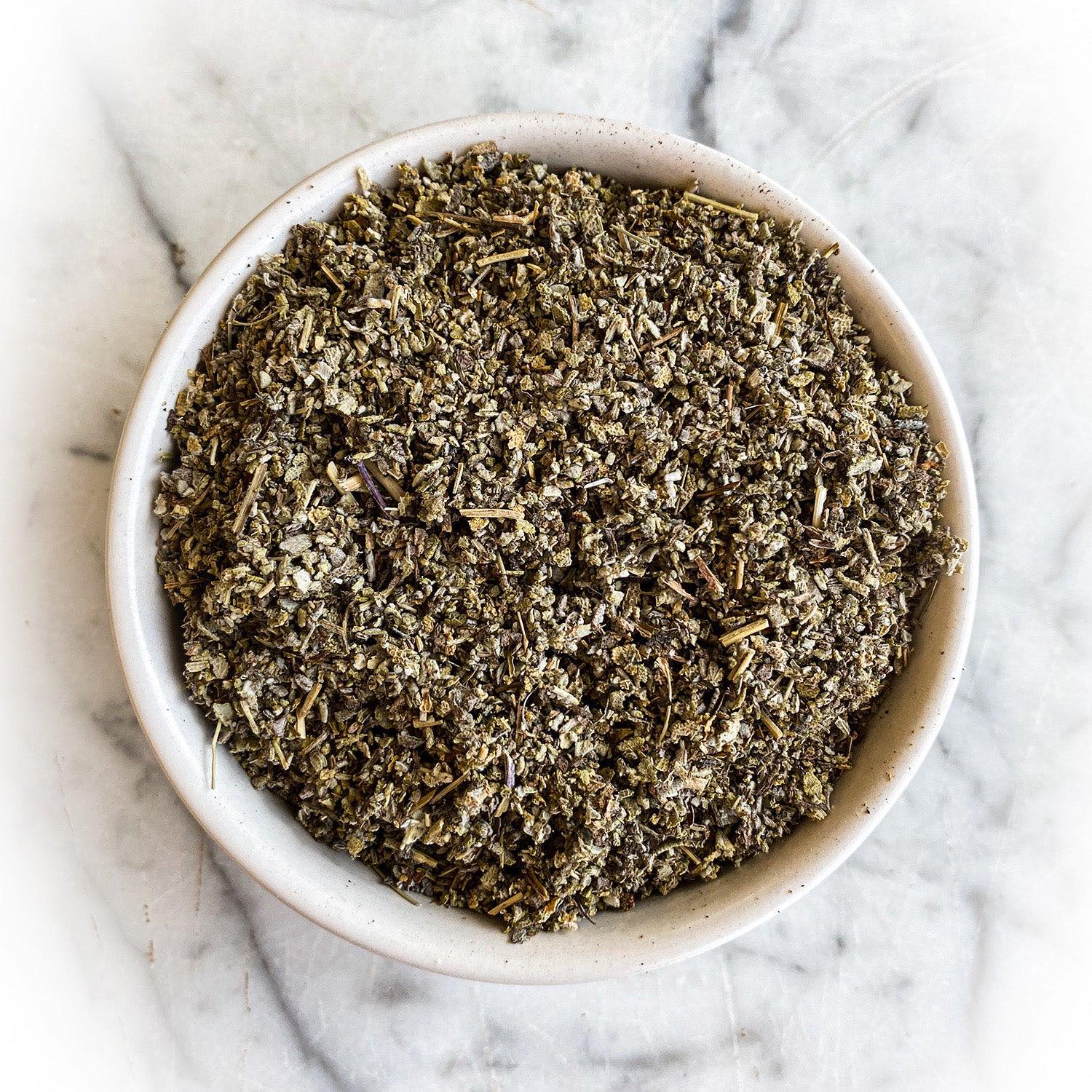 Dried and cut sage leaves fill a white bowl on a white marble table.