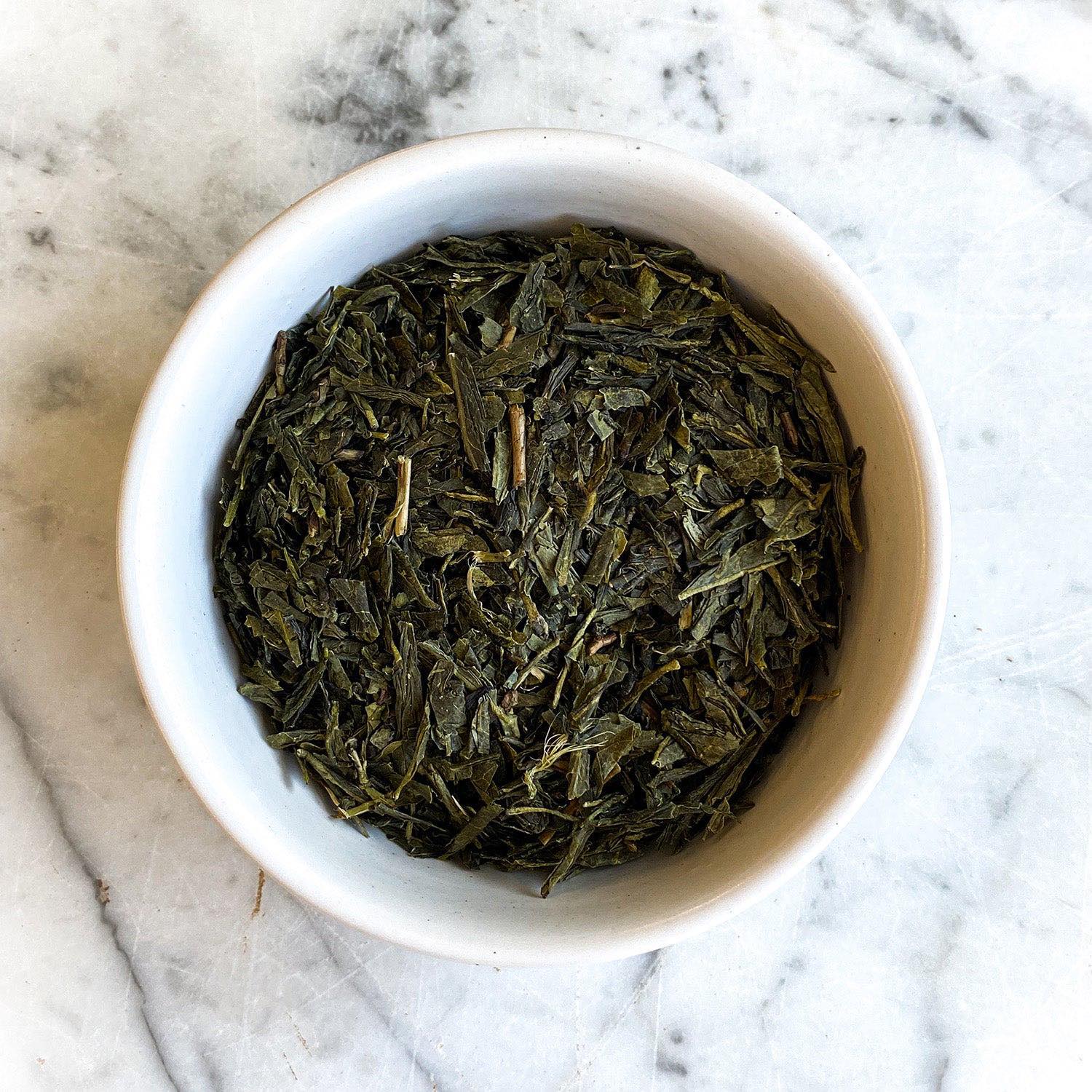 Dried sencha green tea is displayed in a white bowl on a white marble countertop.