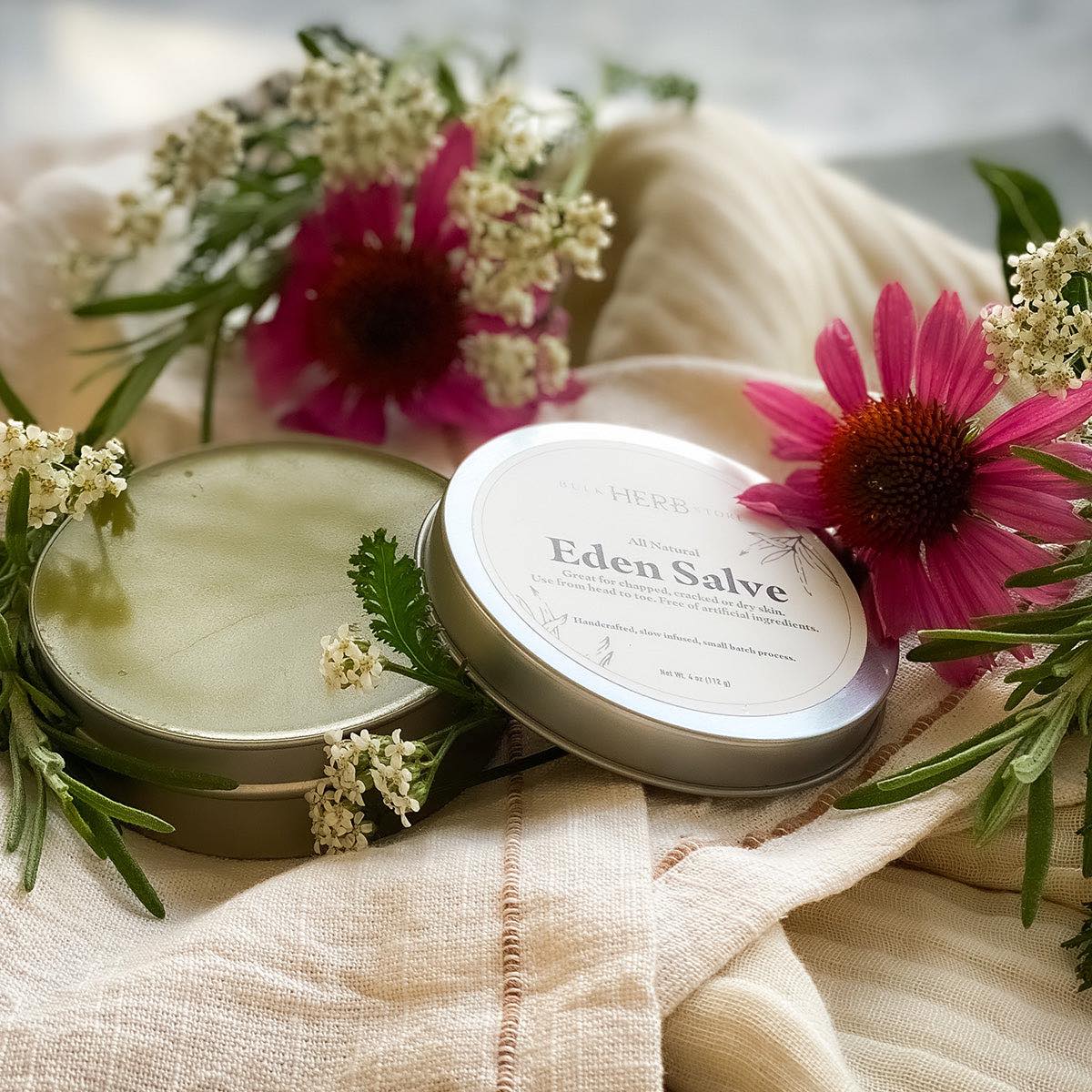 An open tin of Eden Salve rests on a linen cloth with flowers and herbs.