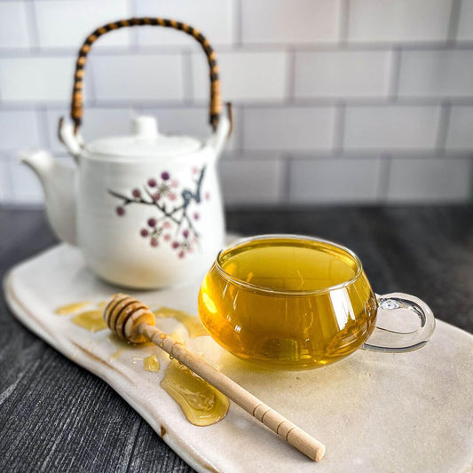 A brewed cup of Fast Asleep Tea rests on a white ceramic surface with a honey wand and white tea pot.