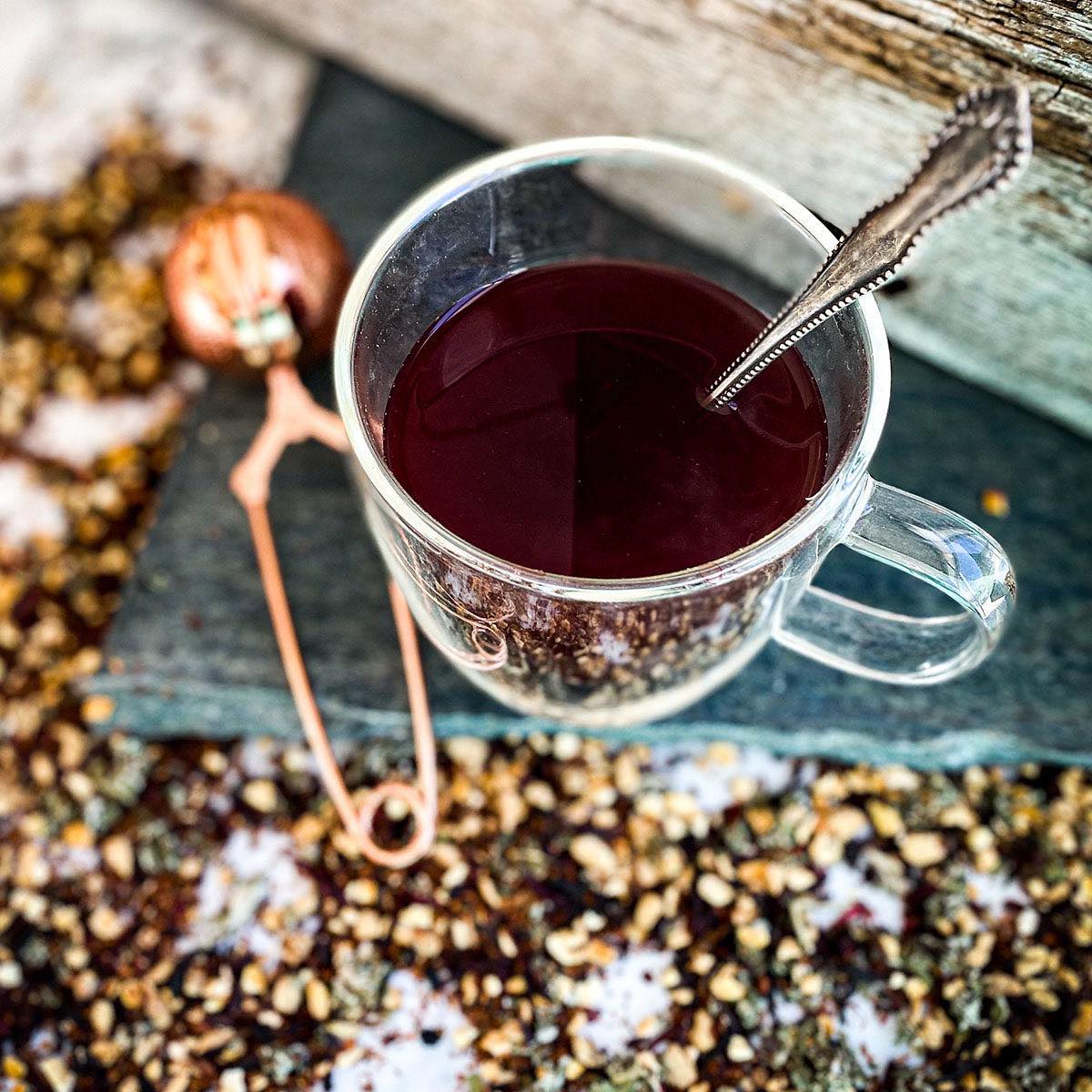 A brewed mug of Herban C tea rests on a stone surface surrounded by dried loose tea.