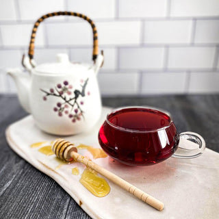 Hibiscus flower tea in a clear mug with a white tea pot in the background and a honey wand