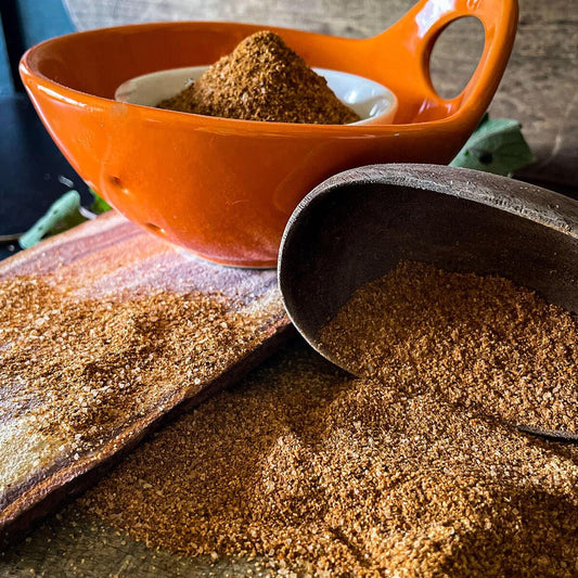 A wooden spoon tips over spilling the jerk seasoning on the counter. An orange dish in the background holds a white bowl of Jamaican Jerk Seasoning powder.