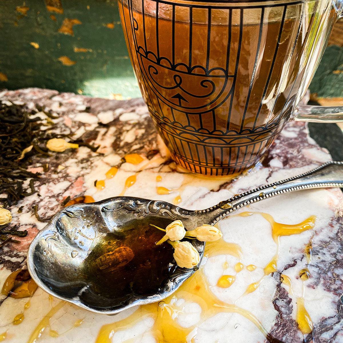A cup of brewed Jasmine Green Tea is in the background. A spoon with honey is in the foreground.