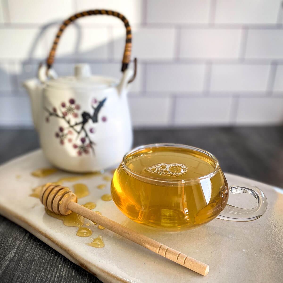 Jasmine Green tea brewed in a clear glass mug with a honey wand and a white tea pot in the background.