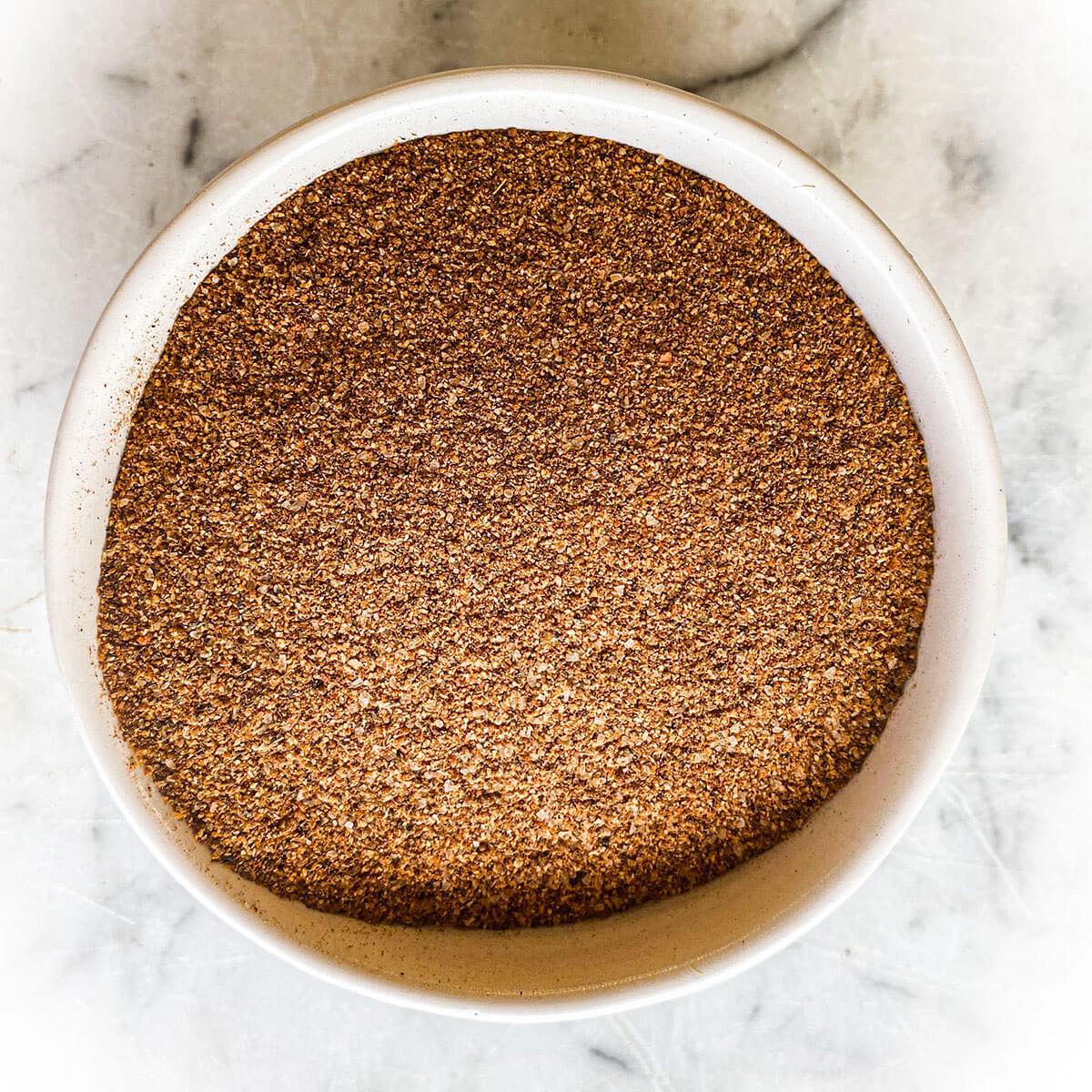 A white bowl filled with Jamaican Jerk Seasoning Powder sits on a white marble counter.