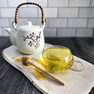 A glass tea cup filled with brewed red clover tea sits on a white ceramic surface with a white tea pot and honey wand.