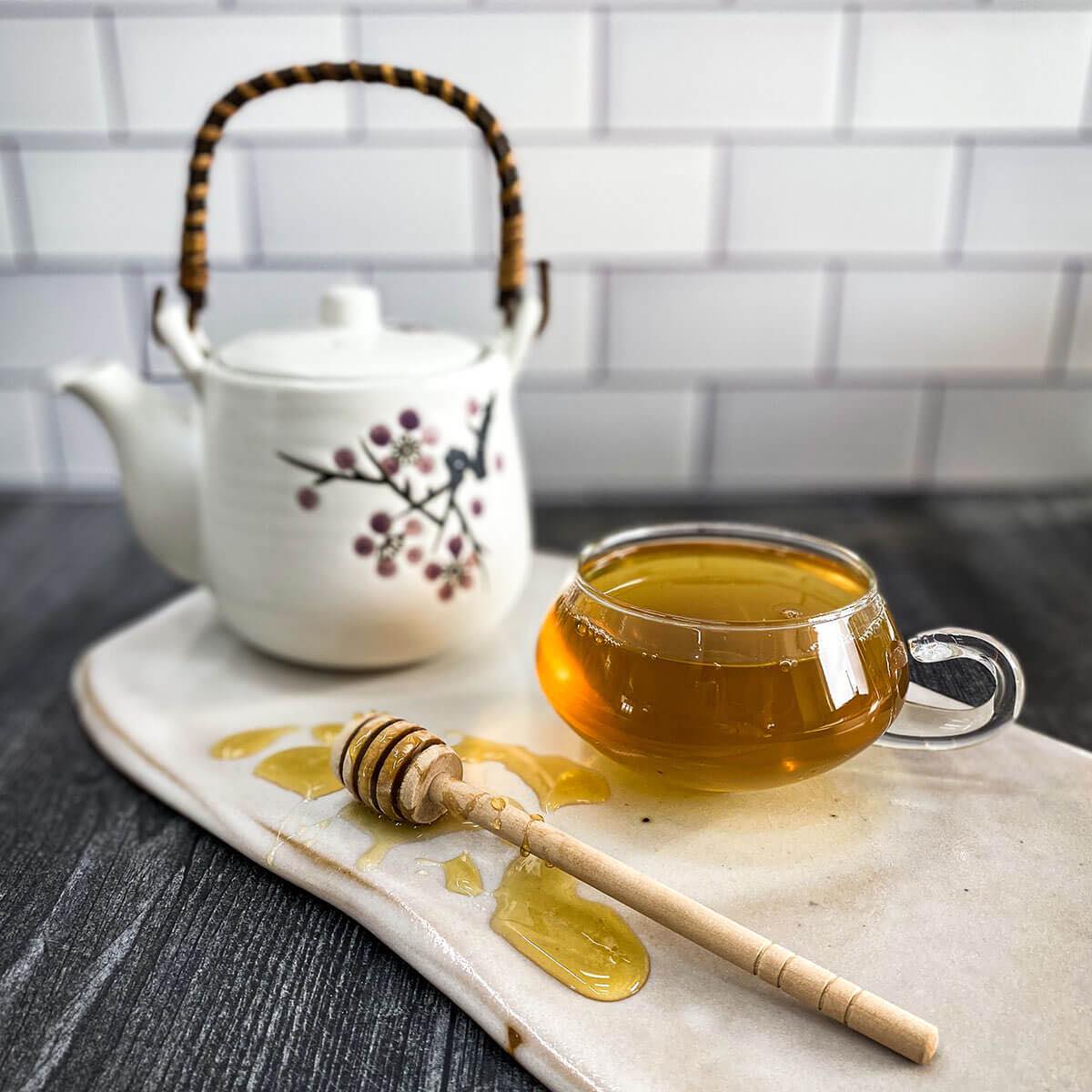 A clear cup of red raspberry leaf tea brew in the foreground with a white tea pot in the background.