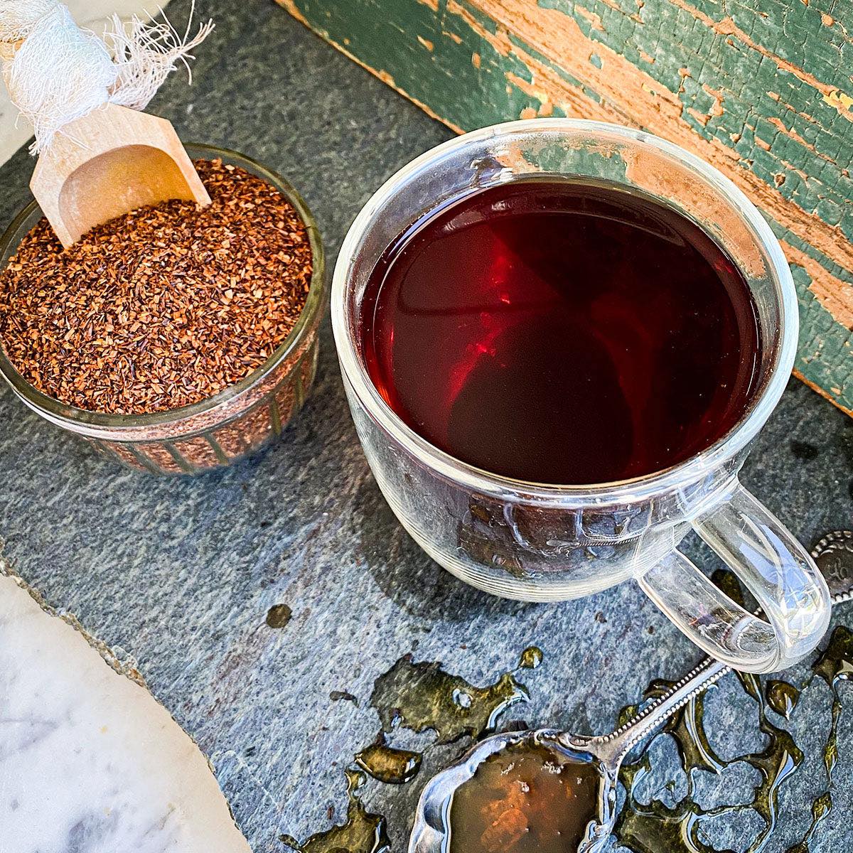 A brewed cup of red rooibos is flanked by a spoon dripping with honey and a jar filled with the dried tea and a scoop.
