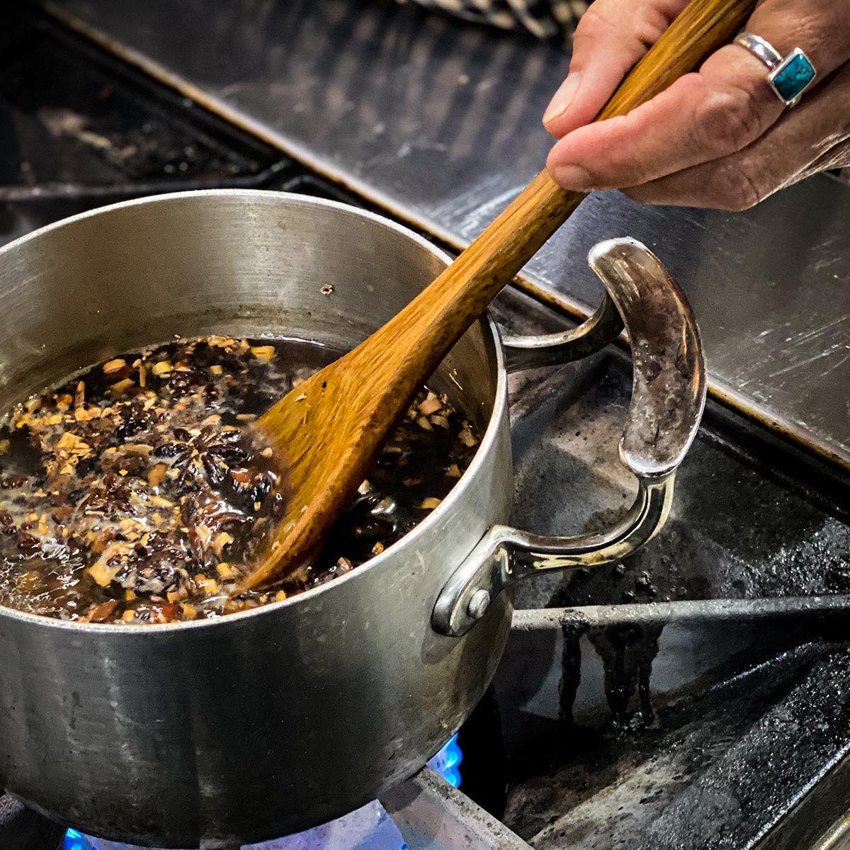 A pot over a stove top contains the sarsaparilla mix recipe and is being stirred by a wooden spoon.