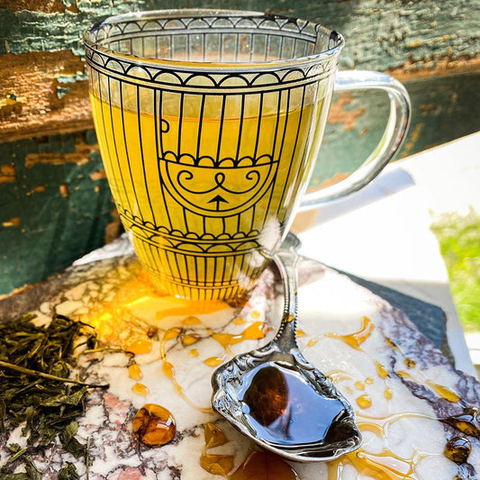Sencha green tea brewed in a decorative clear glass mug with a spoon of honey in the foreground.