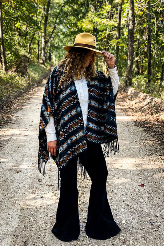 Girl standing on dirt road wearing black fringe poncho with aztec design in colors orange blue and white