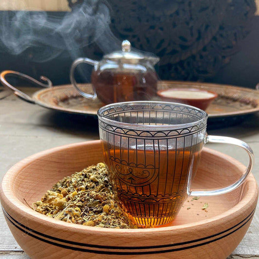 Steam coming from a glass teacup in a wood bowl with loose tea on the side. A glass teapot and tea tray are in the background.