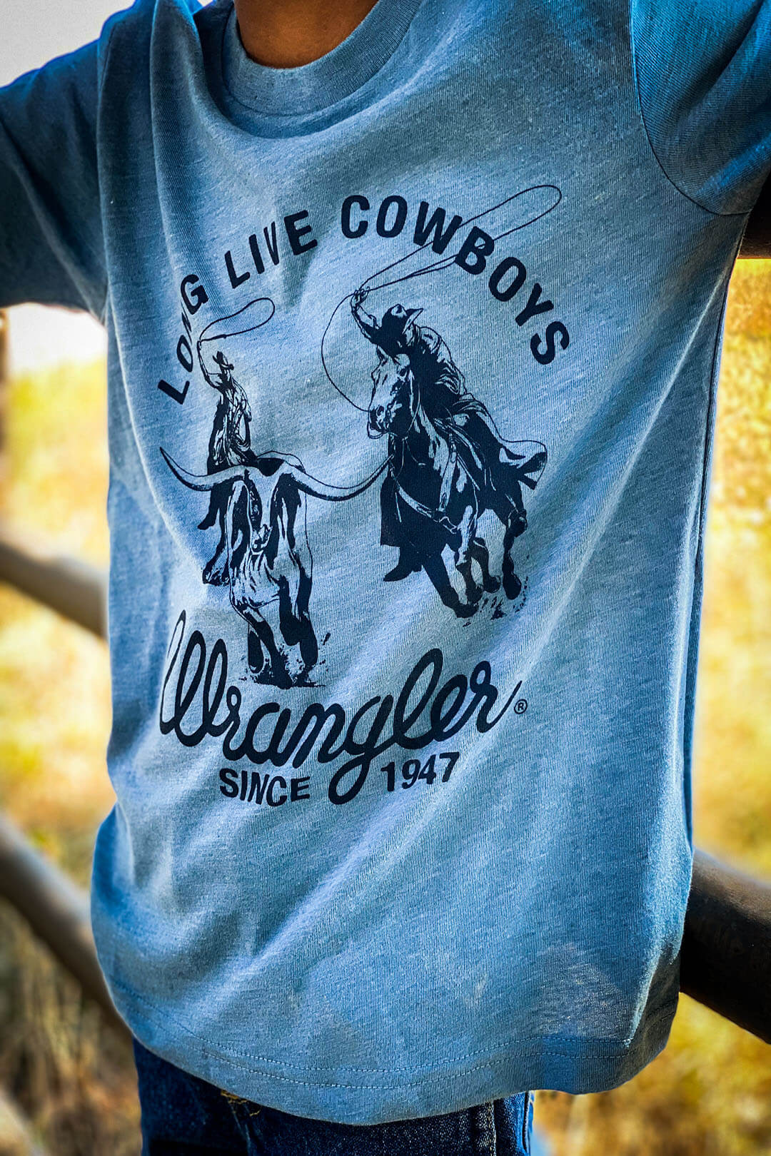 Boy wearing light blue t-shirt with navy blue writing that says long live cowboys wrangler since 1947 cowboys on horses roping bull