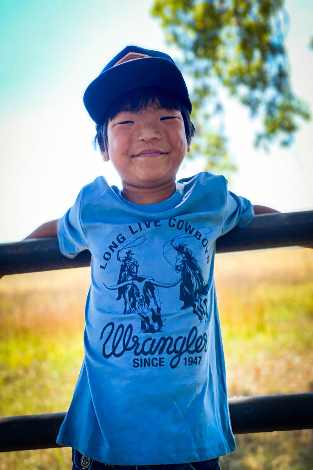 Boy wearing navy ball cap light blue t-shirt with navy blue writing that says long live cowboys wrangler since 1947 cowboys on horses roping bull