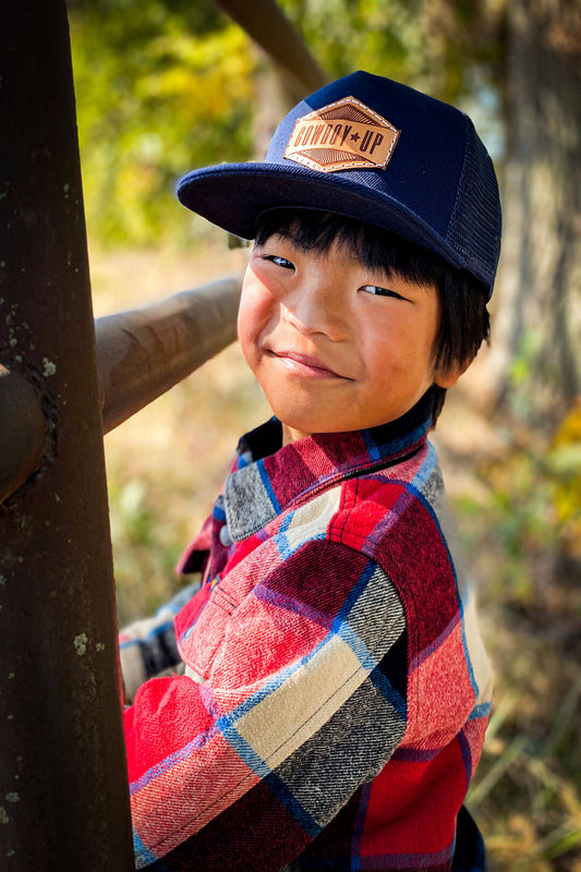 Boy on pipe fence with Wrangler red blue cream black jacket wearing navy ball cap with words Cowboy Up on front of hat