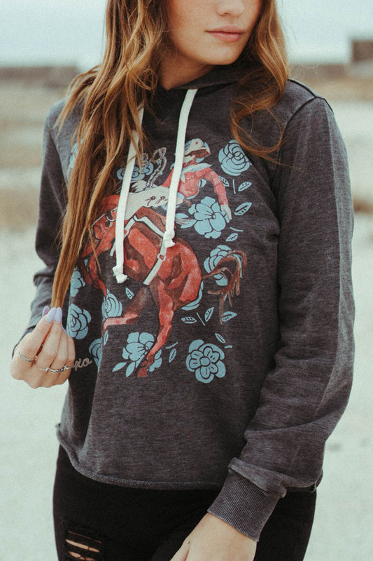 Woman wearing the Bronc Rider Rose Graphic Hoodie with drawstring.  The hoodie is made in Texas with crafted artisan work  Features bronc rider with roses all around. 