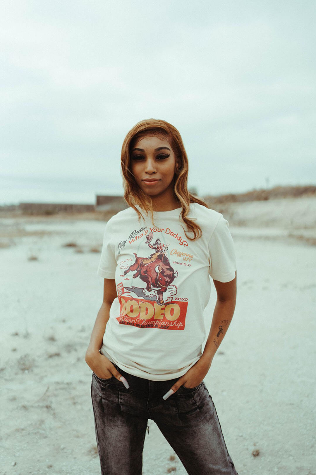 Girl standing with her hands in pocket modeling the XOXO brand Rodeo Tee.  The shirt features a bullrider on a bull. 
