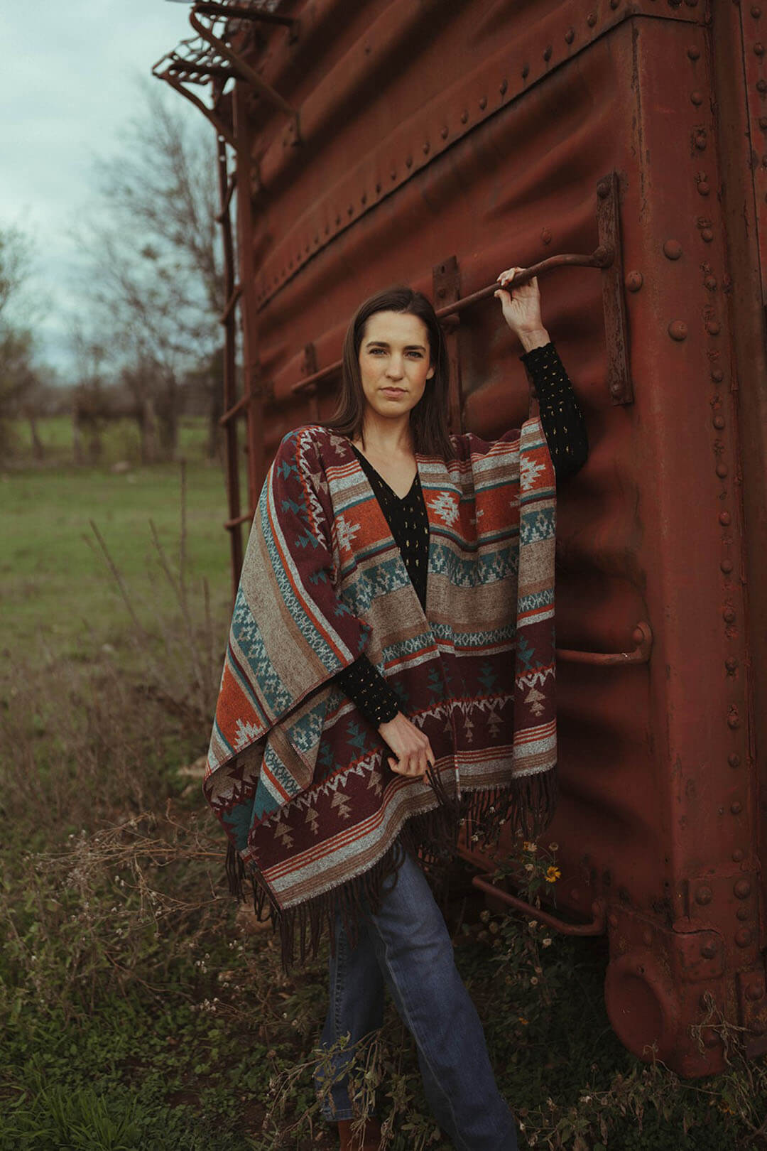 Woman standing beside an old train car modeling the cripple creek aztec poncho.  The poncho has fringe detail on bottom. 