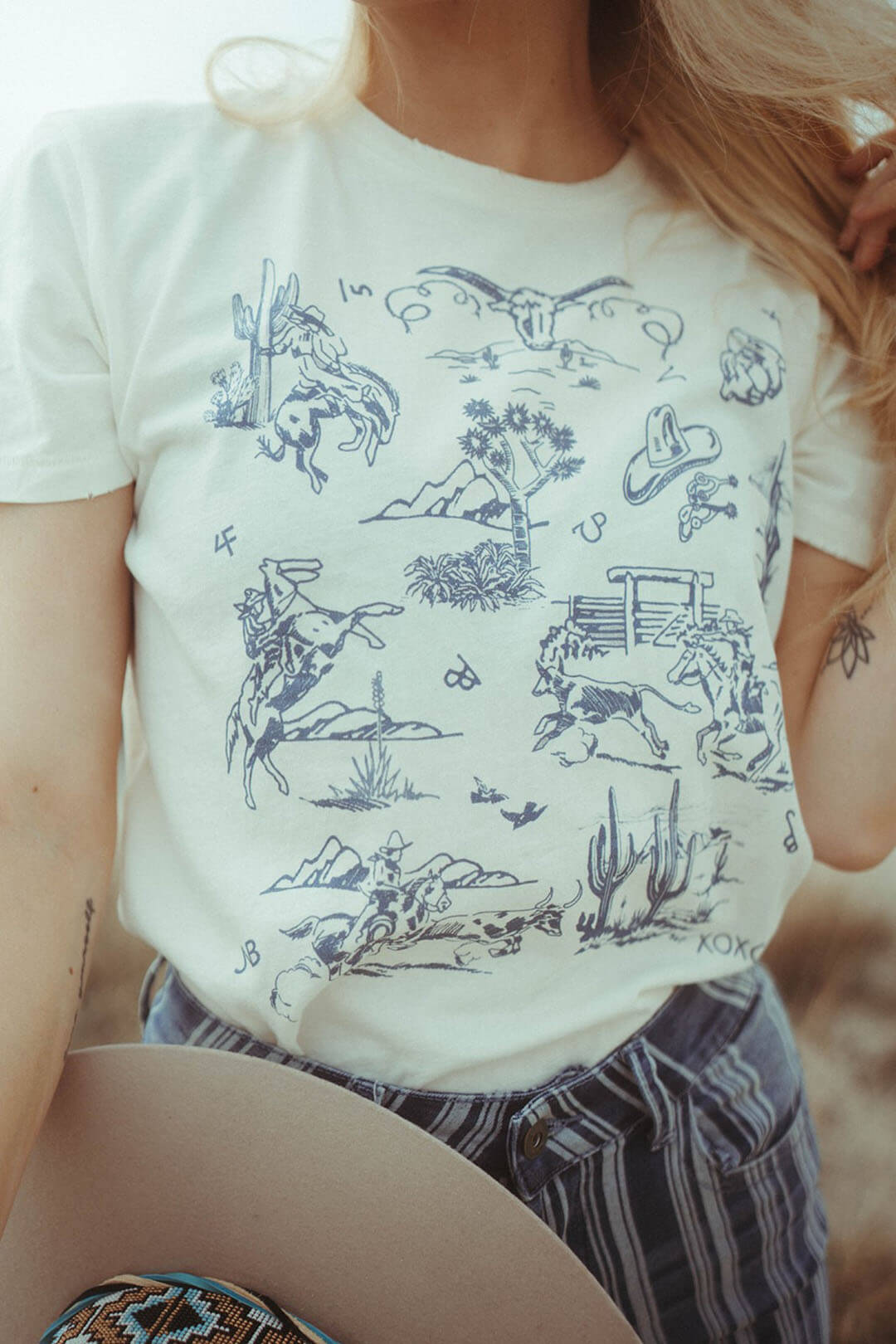Close up image of the Western Toile Denim Graphic Tee from XOXO art & comanpy featuring western images throughout from cactus, cowboys, and cowskulls.