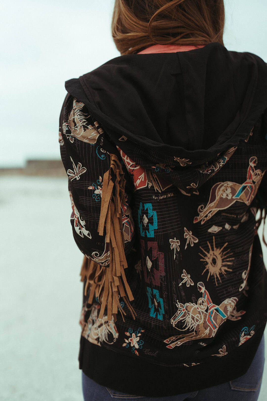 Woman modeling the back side of the double d ranchwear ladies dragonfly riders hooded jacket.  The jacket features indians on horseback with aztec design throughout and the arms have fringe on the bottom of each side. 
