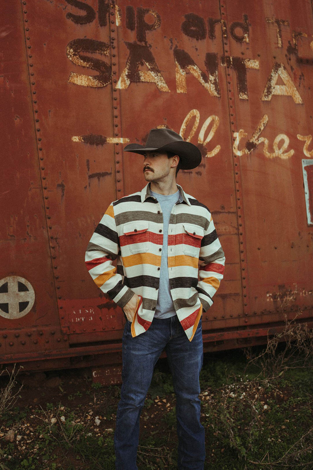Man standing near an old train wearing the driftwood shirt that can be used as a jacket.  Button up jacket with a striped look with yellow, red, gray, and black stripes. 