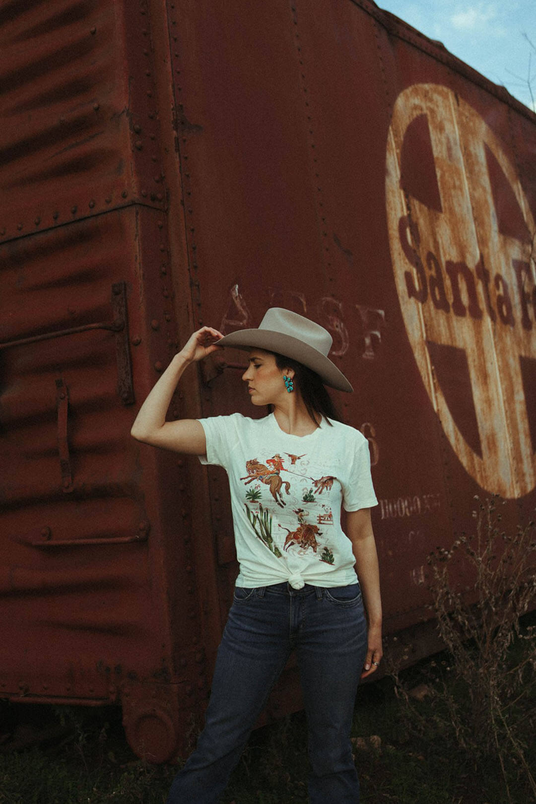 Woman standing near train car wearing the Grandpas Wallpaper Graphic Tee by XOXO.  Western scene on shirt