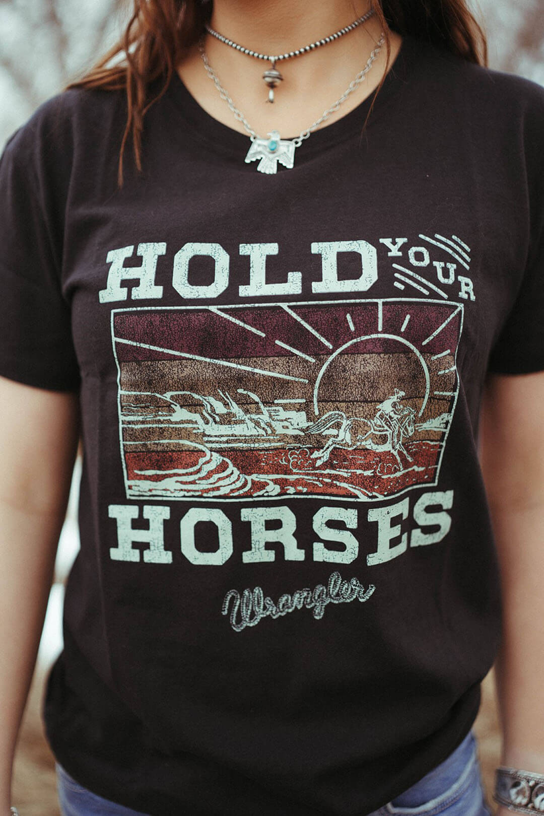 Woman modeling the "Hold Your Horses" graphic tee.  The shirt is black in color with a colorful western scene of a cowboy riding along side of the sun.