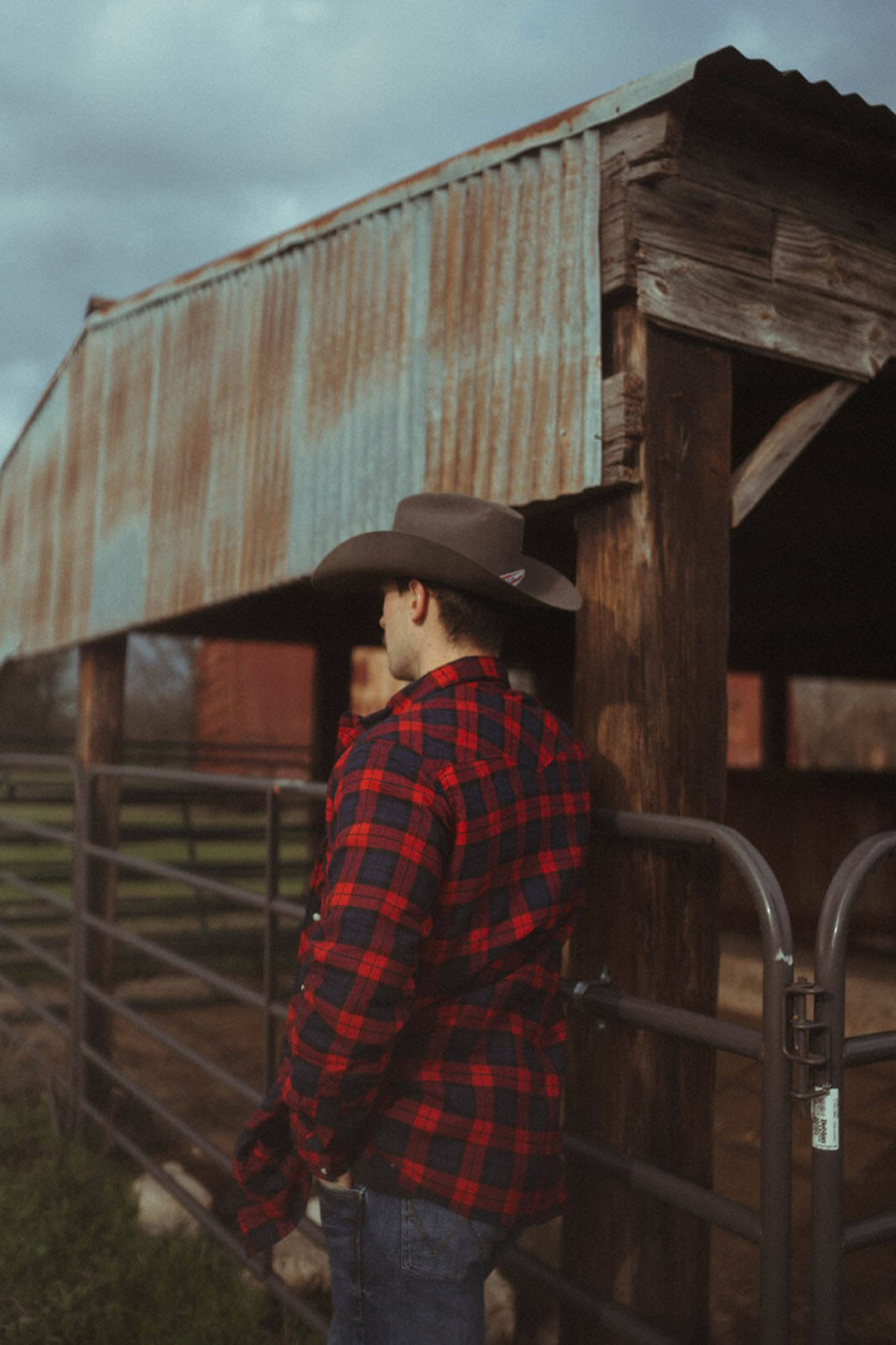 Man leaned up against gate showing the back of the quilted lined flannel wrangler button up jacket.  The shirt features a black/red plaid look.