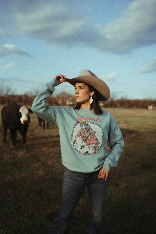Woman standing in field wearing the rodeo resist pullover by the XOXO shirt company.  The pullover features a cowboy on a buckin bronco and says "Rodeo"