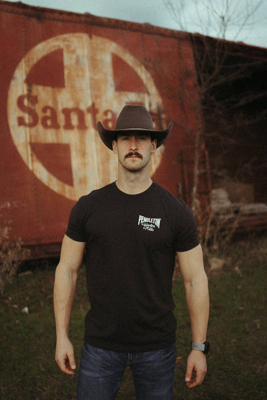 Man standing next to an old train car in field wearing pendleton black graphic tee with the word "Woolen Mills" written under the pendleton logo.