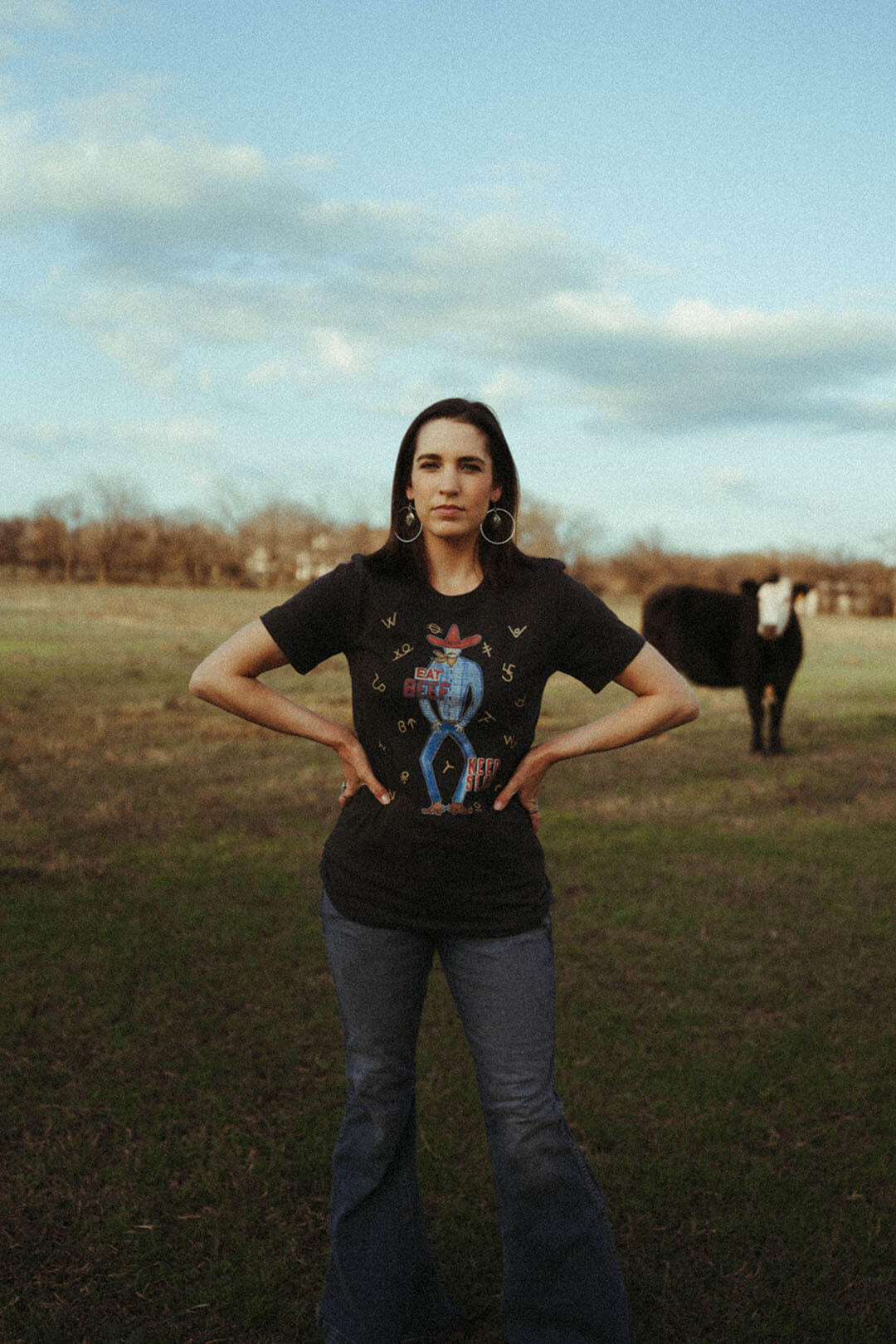 Woman standing in field with a cow behind her modeling the XOXO art & company graphic tee shirt. 