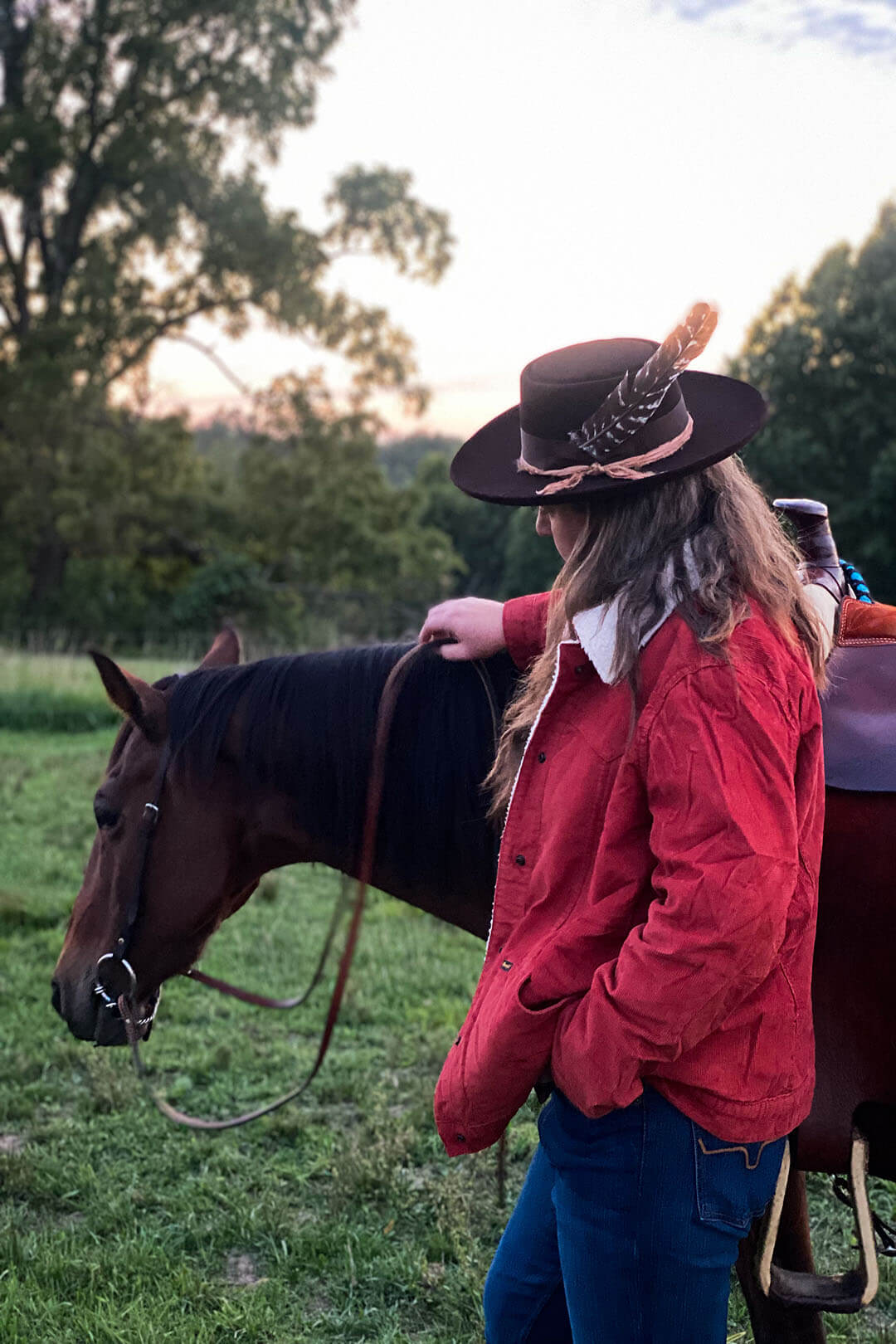Woman holding reins of horse wearing brown hat with feather and rust colored sherpa lined Wrangler brand jacket