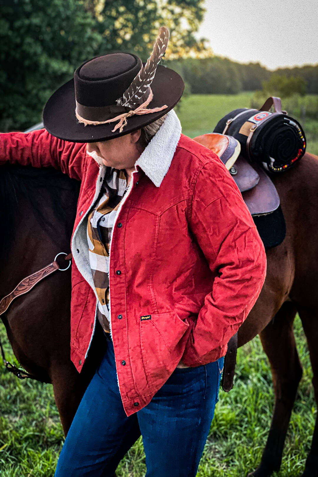 Woman next to saddled horse with throw carrier on back of horse behind saddle wearing rust colored Wrangler insulated jacket sherpa lining