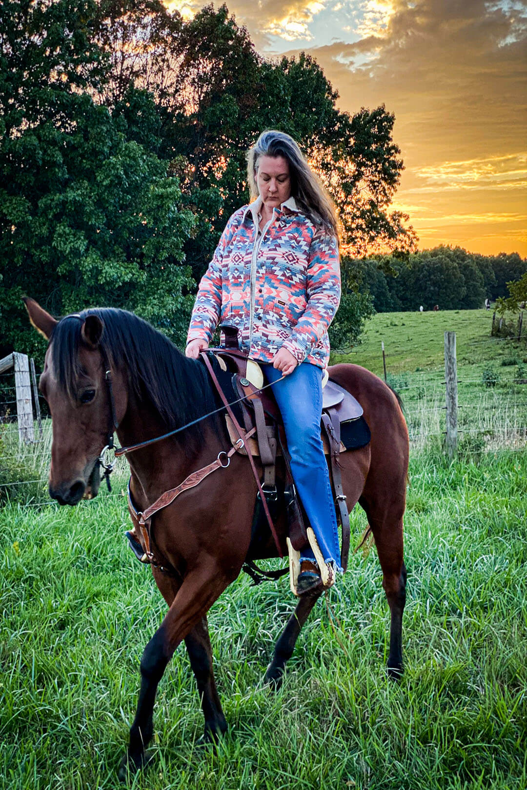 Woman in insulated sherpa aztec jacket, riding horse with sunset behind her.  Jacket southwest coloring, aztec symbols in blue pink red tones