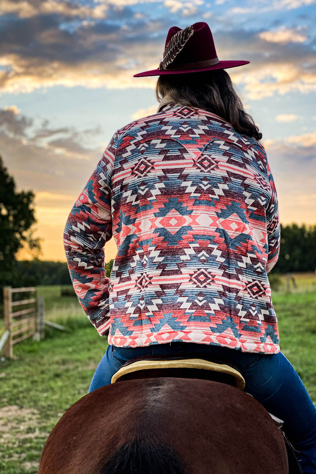 Sherpa insulated jacket southwestern aztec print tones pink, red blue layered throughout entire jacket. Woman on horse wearing burgundy hat with feather stuck in brim