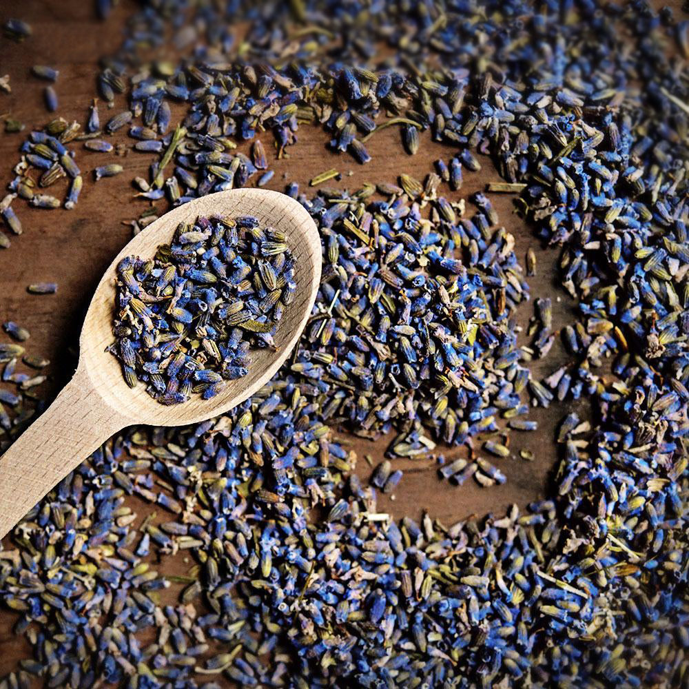 A wooden spoon with Lavender flowers, with more lavender flowers, swirled on a darker wooden surface.