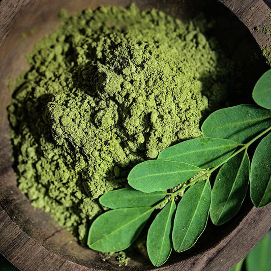 A close-up of the top-view of Green Moringa powder in a dark wood bowl with fresh moringa leaves.