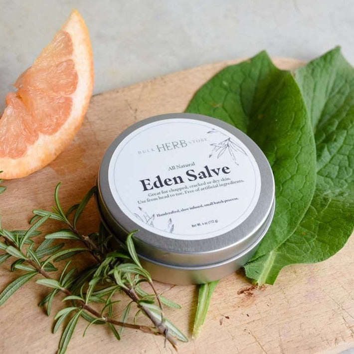 A tin of Eden Salve rests on a wooden cutting board with herbs.