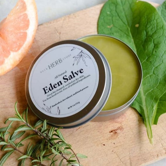An open tin of Eden Salve rests on a wooden board with herbs and a slice of citrus.