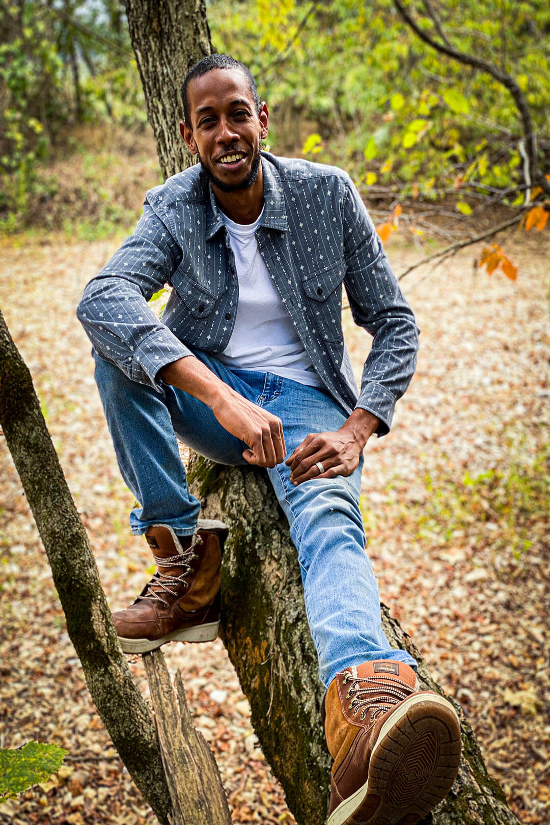 Man wearing corduroy long sleeved shirt gray with small white diamond and line pattern button up front with front pockets sitting in tree denim jeans
