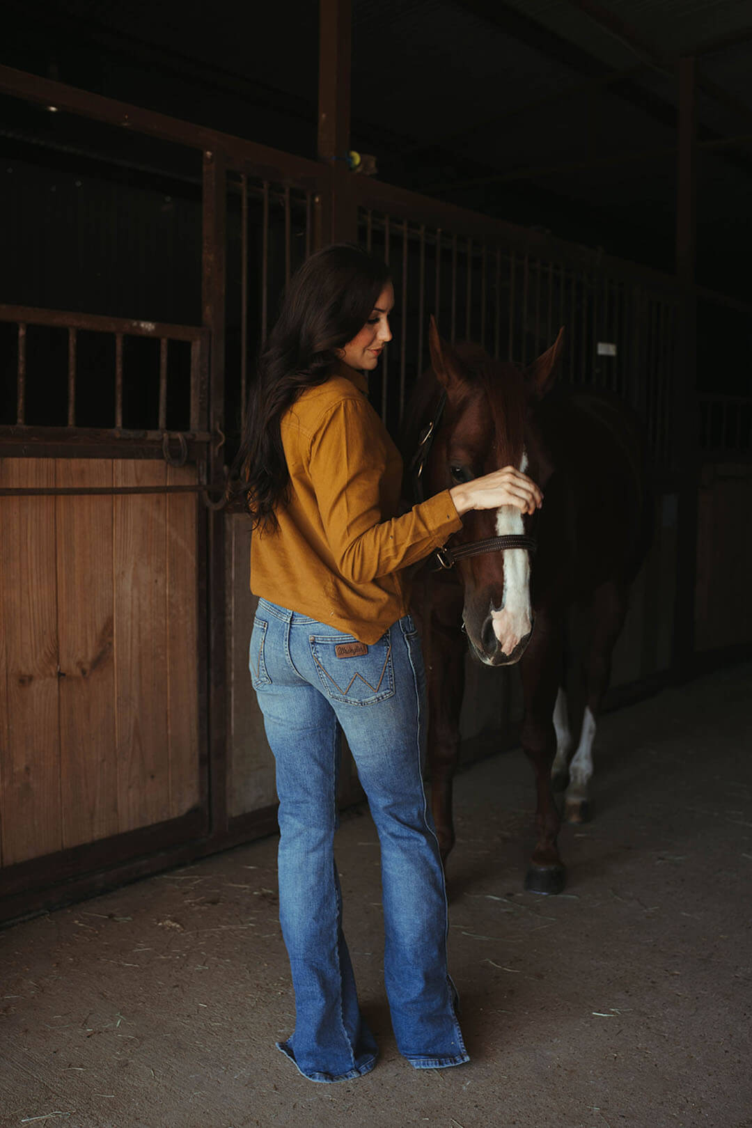 Woman modeling full view of the wrangler retro mid rise bootcut jean while standing next to a horse in barn.  The jeans have slits on each side of outer leg