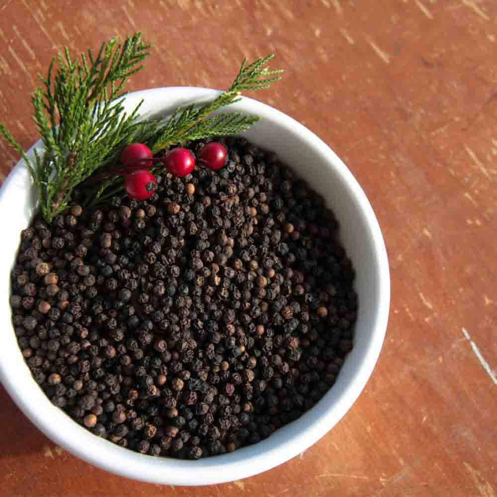 Black Whole Peppercorns with green and red vegetation in a white bowl on a wooden table.