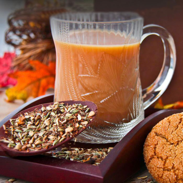A mahogany tray holds a spoon overflowing with loose Gingersnap Tea and a glass mug with tea. Fall décor can be seen and a gingersnap cookie.