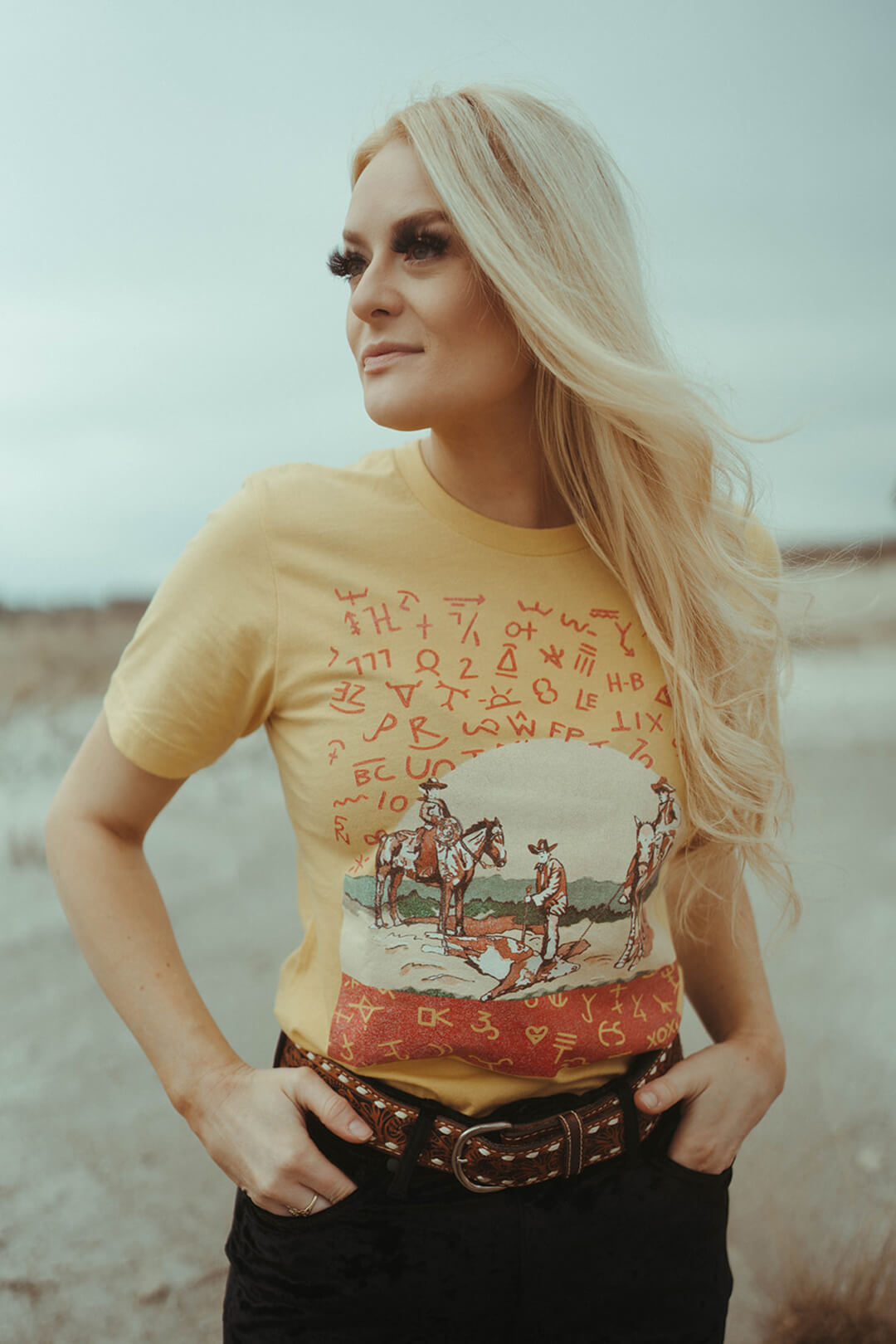 Woman wearing a yellow colored graphic tee featuring picture of cowboys and branding cow.   The shirt also features branding images on the shirt. 
